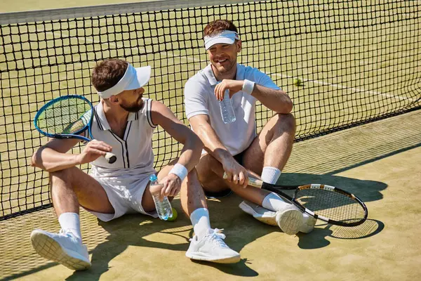 Sahada iki genç adam, eğlenceli bir tenis maçından sonra gülüşmeler ve kahkahalar atıyor..