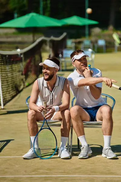 İki genç adam bir tenis kortunda gevşeyip arkadaşça bir maça hazırlanırken yazın tadını çıkarıyor..