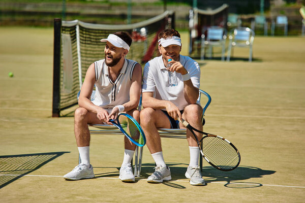 Двое мужчин отдыхают на стульях после игры в теннис, делятся смехом и наслаждаются летним солнцем.