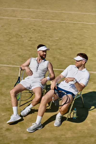 Heyecan verici bir tenis maçından sonra iki adam yaz kıyafetleri içinde gülümseyerek ve kahkaha atarak rahatlıyor..