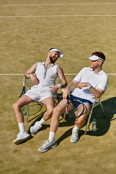 İki erkek tenis sandalyesinde rahatlar, bir yaz maçında gülücüklerini ve gülüşlerini paylaşırlar..