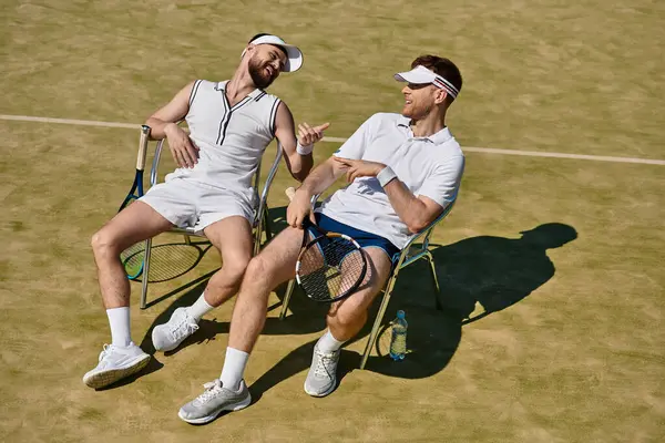 İki yakışıklı adam, yaz tenis kortunda birlikte gülüp rahatlıyor ve eğleniyorlar..
