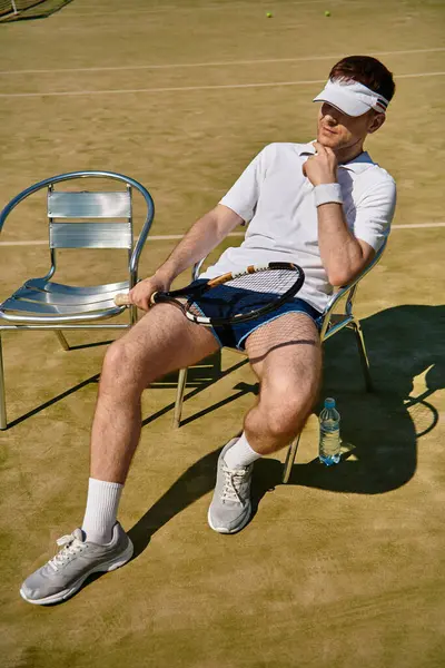 Beyaz spor kıyafetli fit bir adam güneşli bir kortta tenis oynadıktan sonra raketle rahatlıyor..