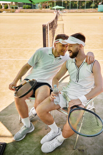 Two men enjoy each others company while resting on the tennis court during summer.