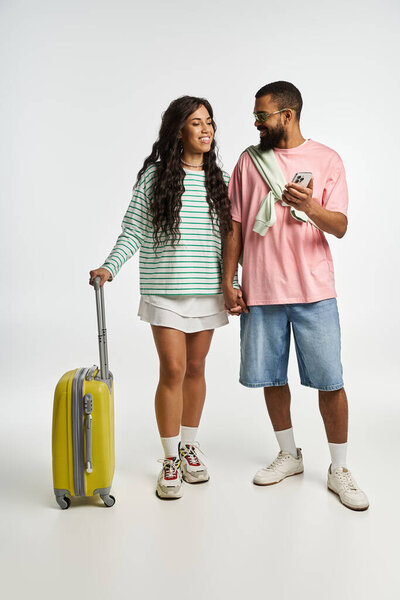 A fashionable couple holds hands and smiles, preparing for a fun trip in a bright studio.