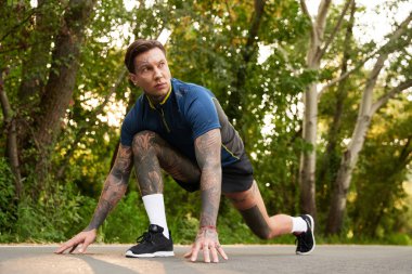 Tarz sahibi genç bir adam, ağaçlarla çevrili manzaralı bir park yolunda egzersiz yapmaya hazırlanıyor..