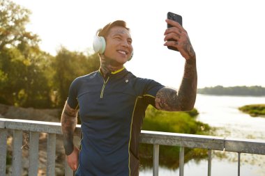 Dövmeli genç bir adam doğa özçekimi için gülümseyerek yürürken duraksıyor..