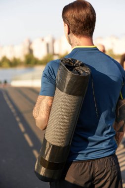 Yakışıklı genç adam nehir boyunca yürüyor, yoga minderi taşıyor, güneşli bir günün tadını çıkarıyor..