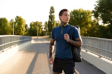 Yakışıklı genç adam güneşli bir yolda yürüyor, atletik stilini ve kendine güvenini gösteriyor..