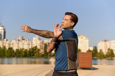 Dövmeli bir genç adam güneşli bir günde suyun kenarında uzanır. Şehir manzaralı..