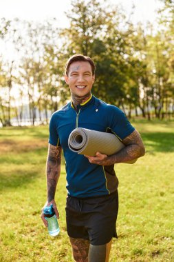 Yakışıklı genç bir adam, elinde yoga minderi ve su şişesiyle güneşli bir parkta geziniyor..