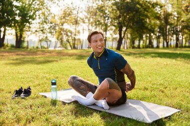 Dövmeleri olan genç bir adam, yeşil ve güneş ışığıyla çevriliyken yoga minderinde geriliyor..