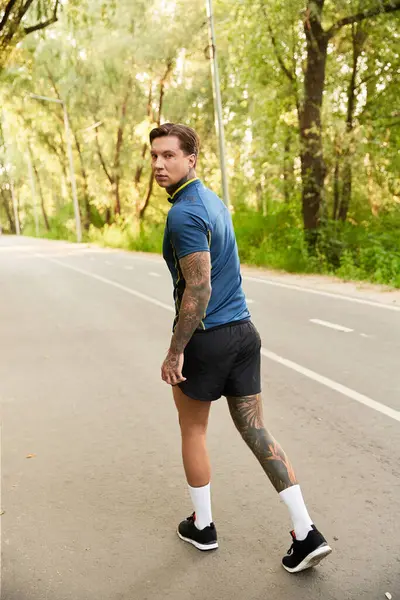 Dövmeleri olan yakışıklı genç adam yemyeşil yemyeşil bir yolda yürüyor.