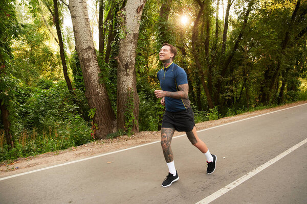 Handsome young man jogs cheerfully along a peaceful road bordered by lush greenery.