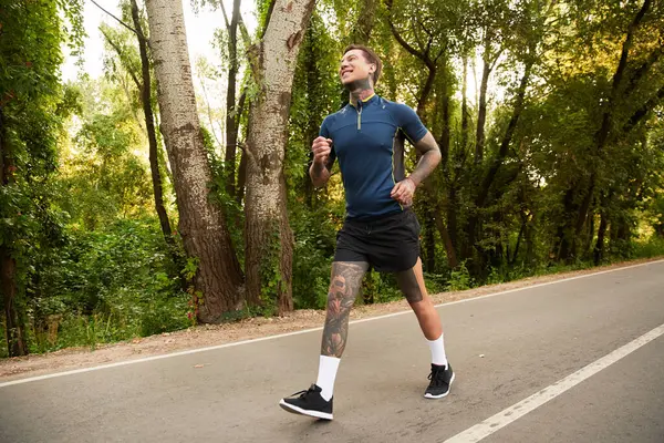 Dövmeleri olan genç bir adam ağaçlarla çevrili manzaralı bir sokakta enerjik bir şekilde koşuyor..