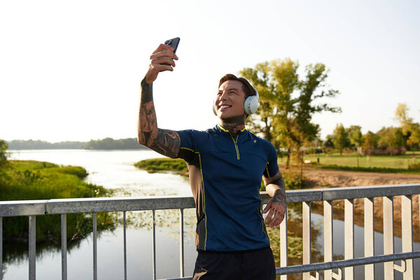 Young man with tattoos walks by the river, smiling and taking a selfie while wearing headphones.