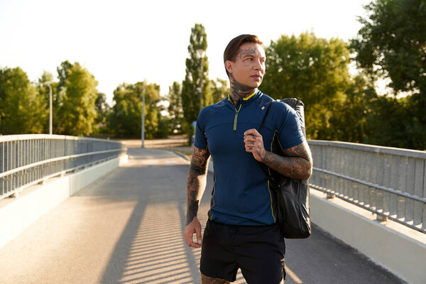 Красивый молодой человек прогуливается по освещенной солнцем дорожке, демонстрируя свой спортивный стиль и уверенность.