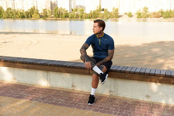 A young man sits quietly on a bench by the river, appreciating the serene surroundings and sunshine.