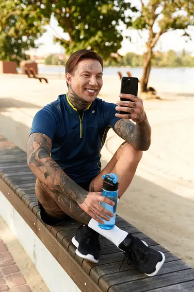 Dövmeli çocuk telefonuna gülümsüyor, güneşli göl kenarında yürüyüş parkurunda mola veriyor..