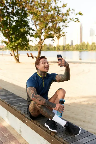 Dövmeli genç adam su kenarında bir bankta oturuyor, gülümsüyor ve güneşin altında selfie çekiyor..