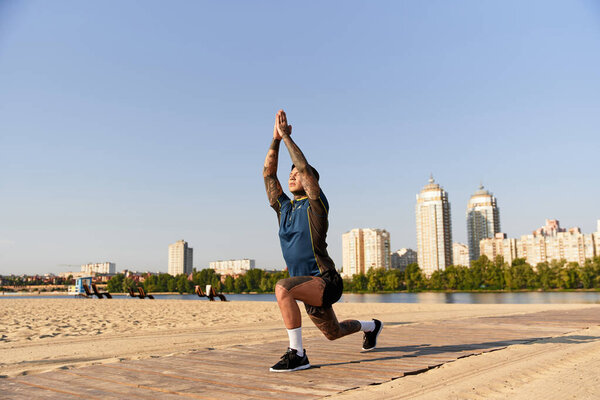 A handsome young man does yoga on the sunny beach with city buildings behind him.