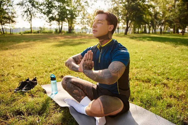 Genç adam yoga minderinde bacak bacak üstüne atmış, huzurlu ve dengeli bir parkta oturuyor..