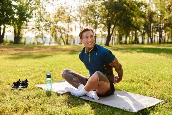 Dövmeleri olan genç bir adam, yeşil ve güneş ışığıyla çevriliyken yoga minderinde geriliyor..