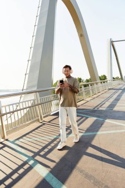 Genç bir adam, güneş ışığı manzarayı güzelleştirirken telefonuyla bir köprüde öylesine geziniyor..