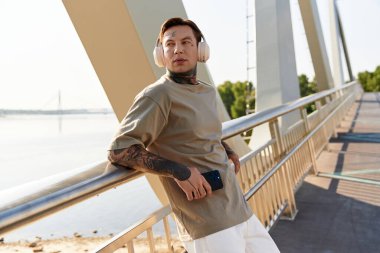 Genç bir adam manzaralı bir köprüde rahatça geziniyor, manzaranın tadını çıkarırken müzik dinliyor..