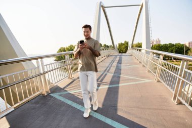 Genç adam köprüde güvenle yürüyor, gülümsüyor ve akıllı telefonuyla kayıyor..