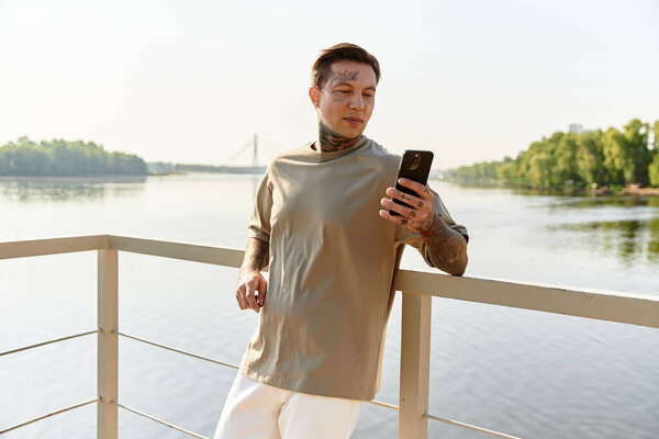 A young man stands on a riverside balcony, admiring the view and using his phone under sunlight.