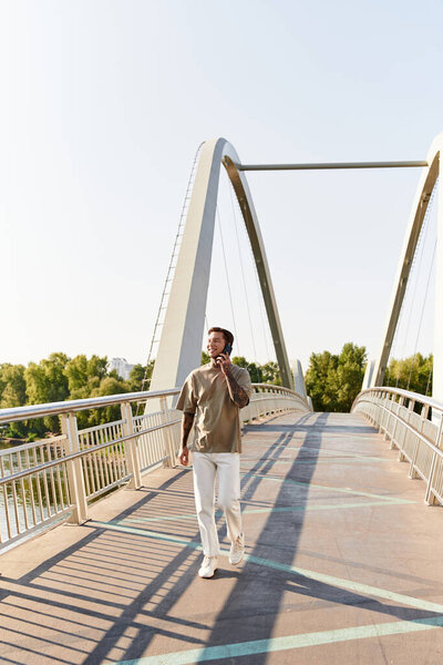Красивый молодой человек неторопливо ходит по пешеходному мосту в ясный день, разговаривая по телефону.