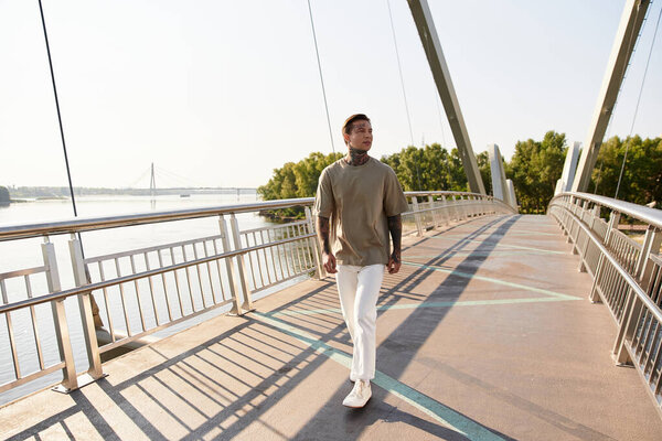Стильный молодой человек уверенно прогуливается по солнечному мосту, окружённый природой и водными видами.