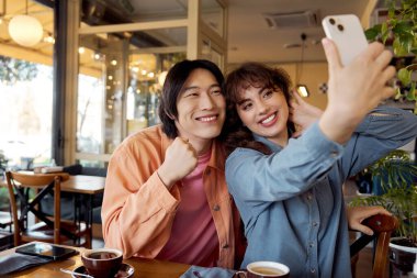 İki arkadaş, sıcak, davetkar bir kafe ortamında selfie çekerken kahkahalarını paylaşıyor..