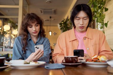 İki genç arkadaş canlı bir kafede yemek yerken telefonlarıyla meşgul oluyorlar..