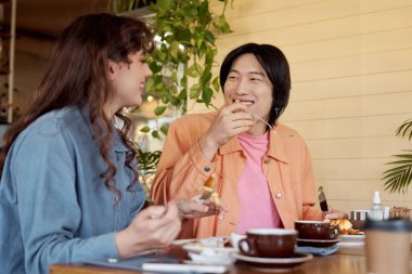 Genç bir Asyalı erkek ve kadın, bir kafede yeşilliklerle çevrili kahvaltıda gülüşürler..