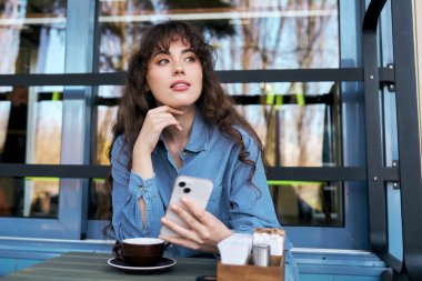 Genç bir kadın hayat dolu bir kafede rahatlıyor, içkisinin tadını çıkarıyor ve akıllı telefon kullanıyor..
