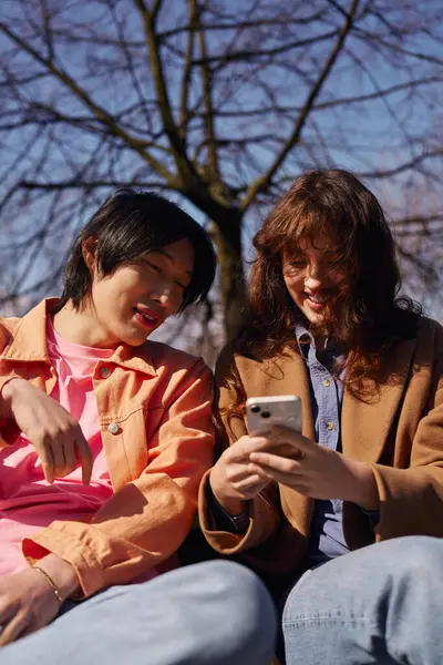 İki genç arkadaş parkta neşeli bir anı paylaşıyor, güneşi emerek ve kahkaha atarak..