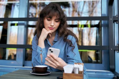 Neşeli bir kafede, genç bir kadın içkisini içiyor ve telefonlarını kontrol ediyor.