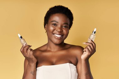 Cildi parlayan genç bir kadın canlı bir stüdyoda cilt serumları tutarken gülümsüyor..