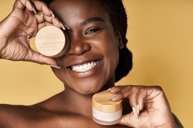 Neşeli genç Afrikalı Amerikalı kadın, parlak bir stüdyoda cilt ürünleri tutarken gülümsüyor..