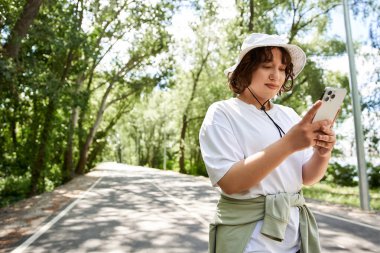 Genç bir kadın güneşli bir yaz gününün tadını çıkarırken dışarıda, ağaçların arasında telefonunu kontrol ediyor..