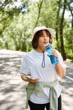 Genç bir kadın rutin egzersizini dışarıda yapar. Susuz kalmadan suyunu yudumlar..