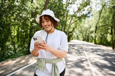 Neşeli bir gülüşü olan genç bir kadın akıllı telefonuyla meşgul, yemyeşil ile çevrili..