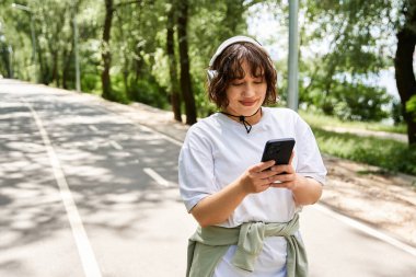 Kulaklıklı genç bir kadın yaz havasının tadını çıkarırken telefonunu doğada kontrol ediyor..