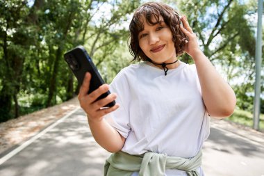Neşeli, kıvırcık saçlı genç bir kadın açık havada güneşli bir yaz gününde müzik dinlerken gülümsüyor..