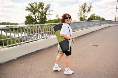 Zinde genç bir kadın, yoga minderini nehir kenarındaki manzaralı bir patikada taşırken gülümser..