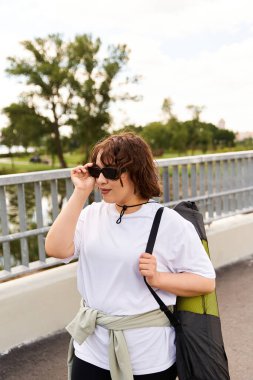Genç kadın dışarıda egzersiz yapıyor, güneş gözlüğü takıyor ve yoga minderi taşırken forma giyiyor..