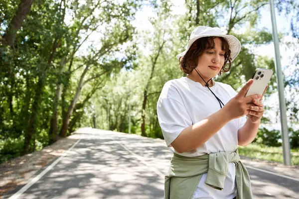 Genç bir kadın güneşli bir yaz gününün tadını çıkarırken dışarıda, ağaçların arasında telefonunu kontrol ediyor..