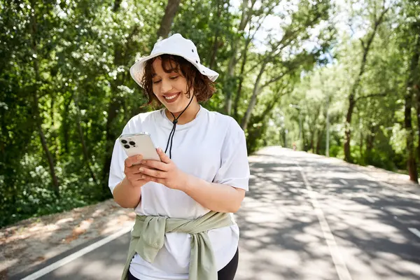 Neşeli bir gülüşü olan genç bir kadın akıllı telefonuyla meşgul, yemyeşil ile çevrili..